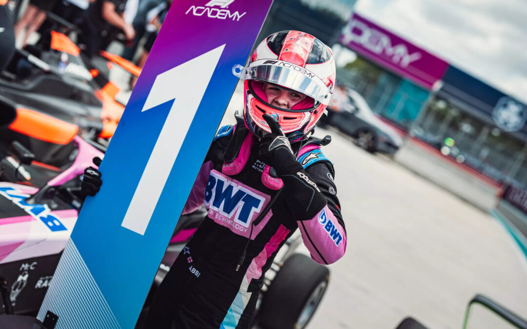 Abbi Pulling next to the P1 sign in the Miami race paddock. She is wearing her Alpine race suit and helmet, and has her index finger pointing up to indicate her first place finish.